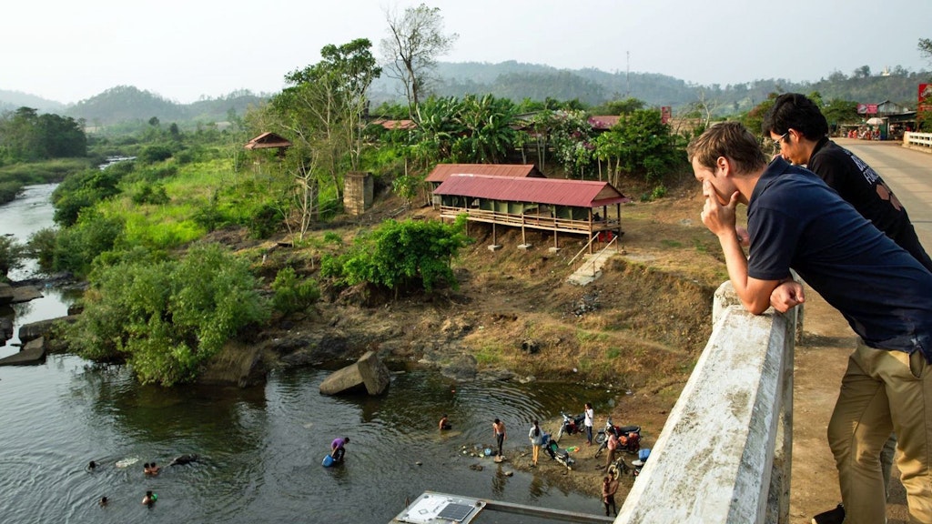 Deux personnes sont appuyées sur le mur d’un pont avec, en contrebas, des personnes s’activant à la mise en place d’un senseur dans la rivière.