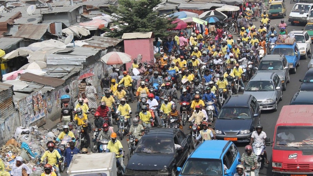 Des voitures et des motos circulent sur une route du Bénin. Des logements de fortune ont été construits le long de la route.