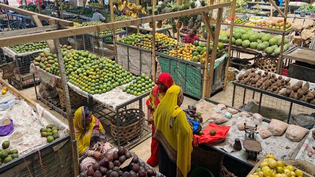 Deux femmes voilées de rouge et de jaune se tiennent devant leur stand de fruits. Des rayons remplis de fruits et légumes les entourent.