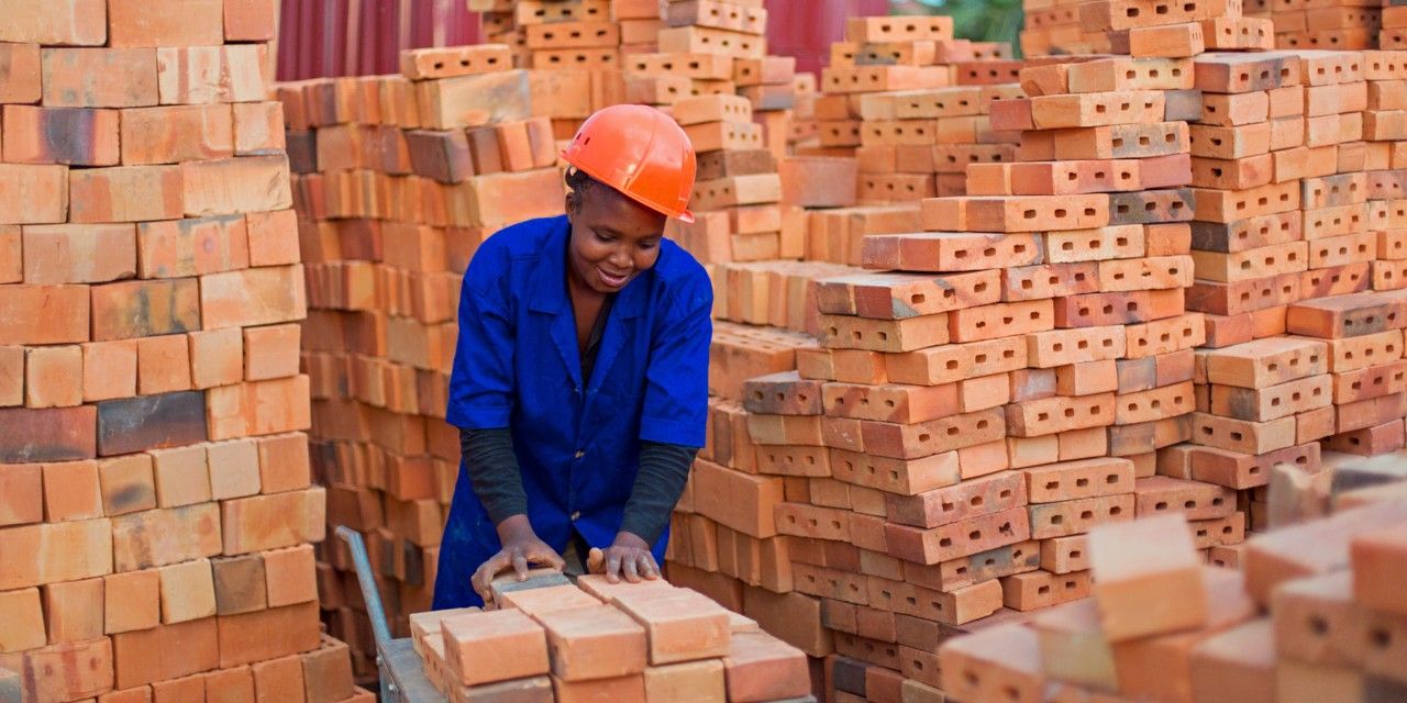 Une femme en bleu de travail, un casque orange sur la tête, dépose des briques sur un chariot.