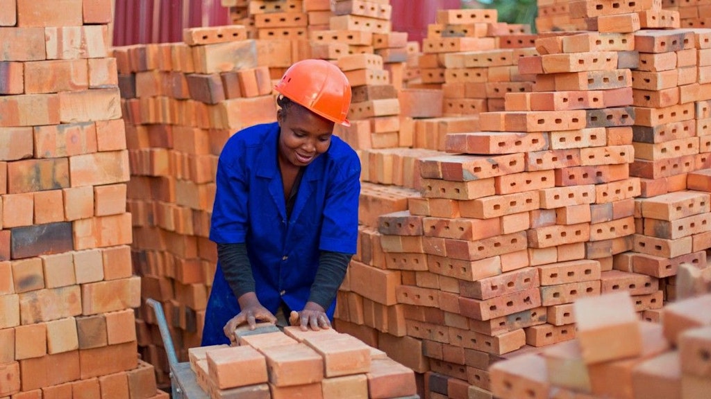 Une femme en bleu de travail, un casque orange sur la tête, dépose des briques sur un chariot.