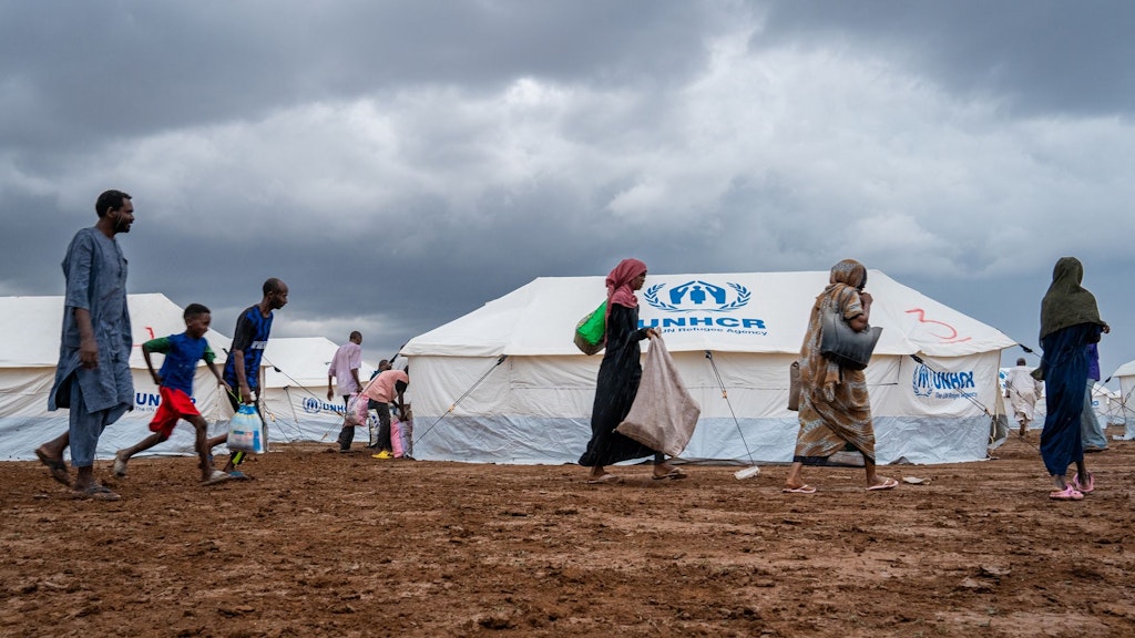 Plusieurs personnes passent à côté d’une tente blanche. Le logo du HCR, imprimé en bleu, est visible sur la tente.