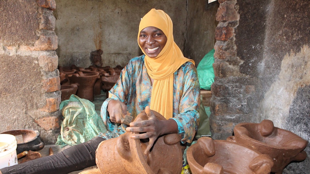 Une femme portant un foulard orange est assise sur le sol au milieu de pots en argile. Elle est en train d’un façonner un nouveau.