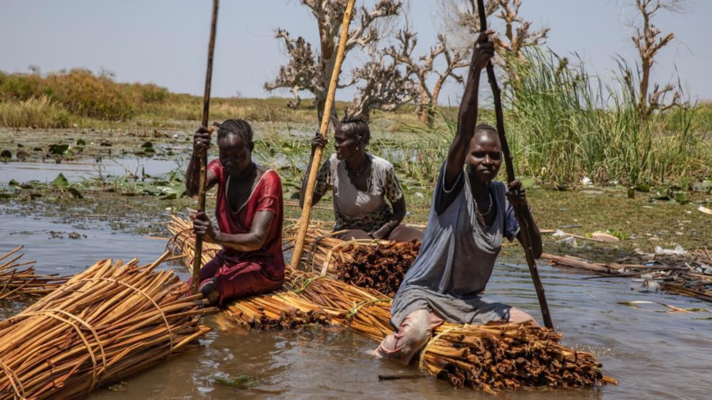 Trois femmes utilisent des fagots de bois comme bateaux pour naviguer dans un paysage inondé.