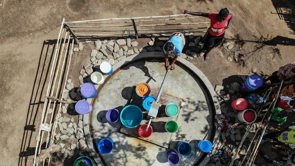 Une pompe à eau est entourée de seaux d’eau colorés. Une femme active la pompe tandis qu’une autre la regarde.