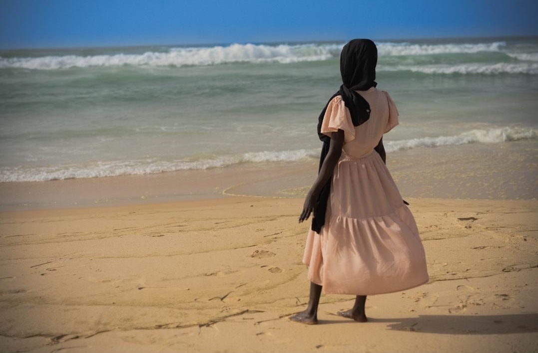 Ein junges Mädchen in einem pastellfarbenen Kleid steht an einem Strand und blickt auf das Meer. Sein Kopf ist mit einem schwarzen Kopftuch bedeckt.