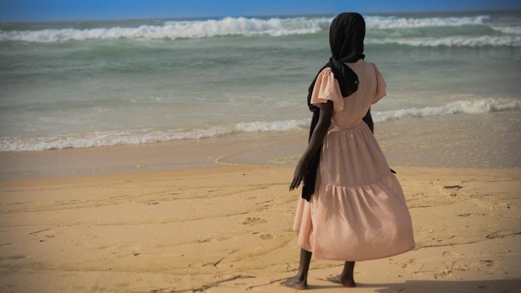 A young girl dressed in a pastel dress is standing on a beach, looking out to sea. Her head is covered with a black scarf.