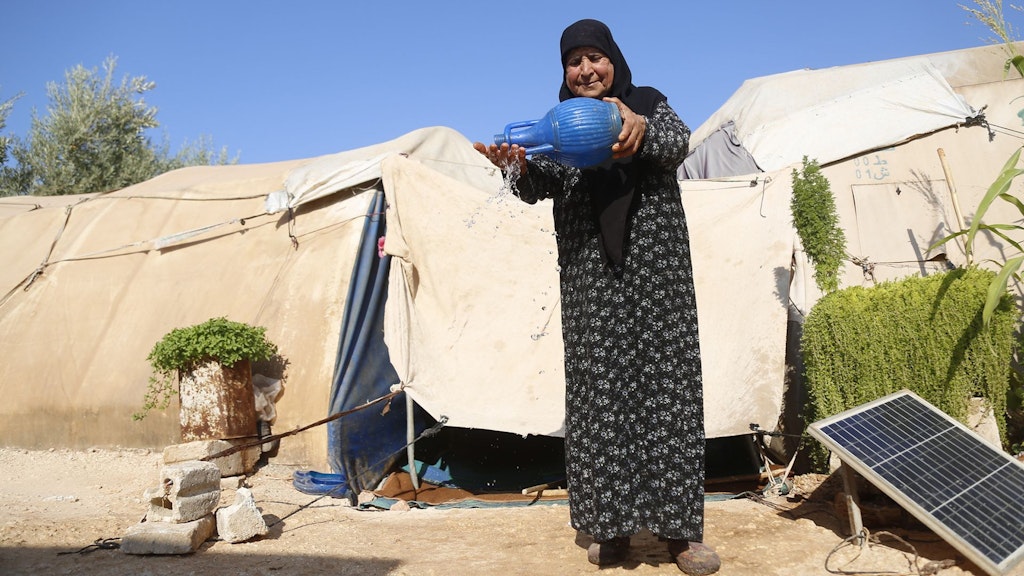 Une femme verse de l’eau à l’aide d’un récipient bleu devant une tente dans un camp de réfugiés, avec de la verdure et un panneau solaire à proximité.