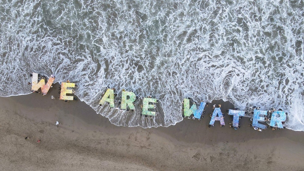 Vue aérienne d’une plage de Bali avec une vague en arrière-plan, où des jeunes déploient des lettres colorées formant le message en anglais «Nous sommes l’eau».