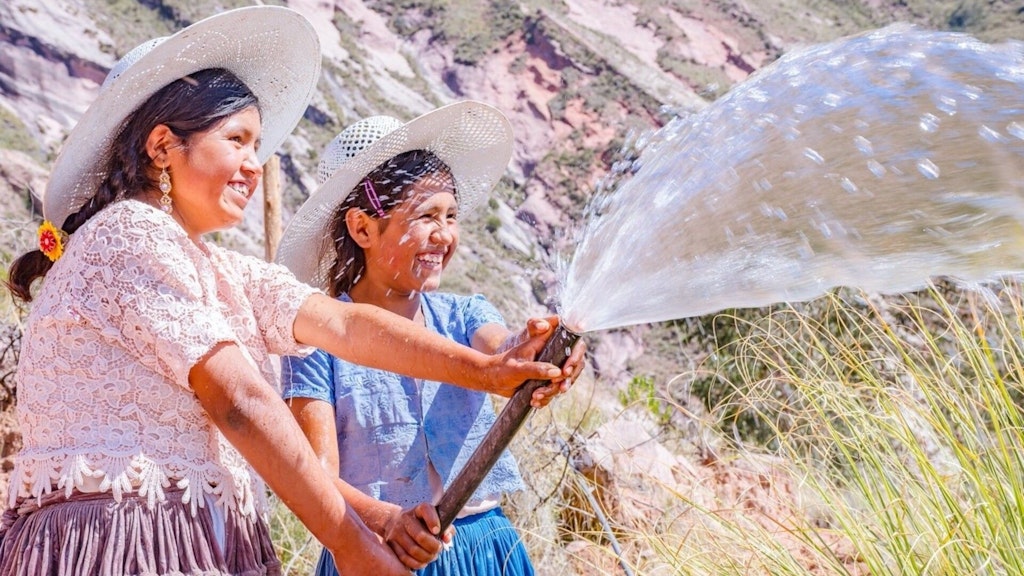 Two smiling girls are holding a water hose together, with fresh water spraying out. They are in a rural, mountainous area with grass and rocky hills in the background.