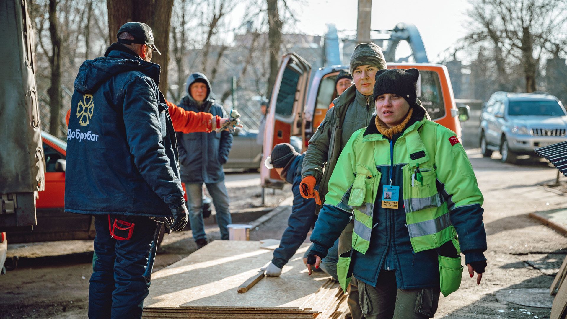 Freiwillige in Winterkleidung tragen Sperrholzplatten bei Hilfsarbeiten im Freien. Im Hintergrund stehen Fahrzeuge und weitere Helfer.