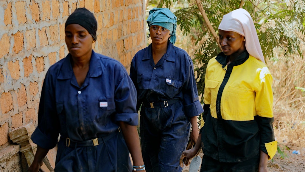 Two young women in dark blue overalls and headscarves walk along a brick wall. Beside them is a woman wearing a yellow jacket and a white headscarf. 