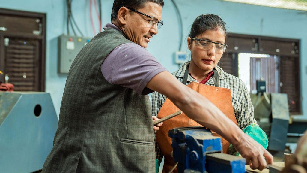Goma Neupane, eine Nepalesin mit zurückgebundenen Haaren, trägt Schutzbrille und steht mit ihrem Chef an der Werkbank. Er zeigt ihr mit einer Feile neue Aufgaben.