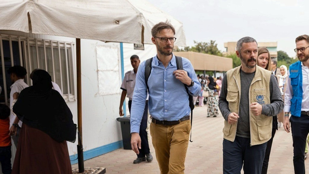 Zwei Männer gehen nebeneinander her. Einer von ihnen trägt ein blaues Hemd und einen Rucksack, der andere eine UNHCR-Weste. Hinter ihnen befinden sich weitere Personen.