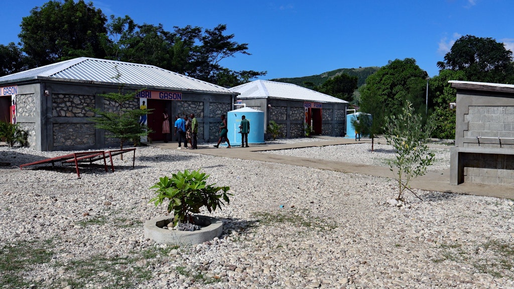 Case in pietra davanti alle quali si trovano serbatoi d’acqua di colore blu. A sinistra si vedono pannelli solari e giovani piante tra le case.  