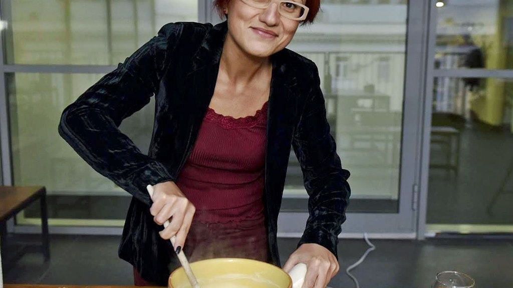 Une femme aux cheveux roux et portant des lunettes remue le fromage dans un caquelon à fondue. 