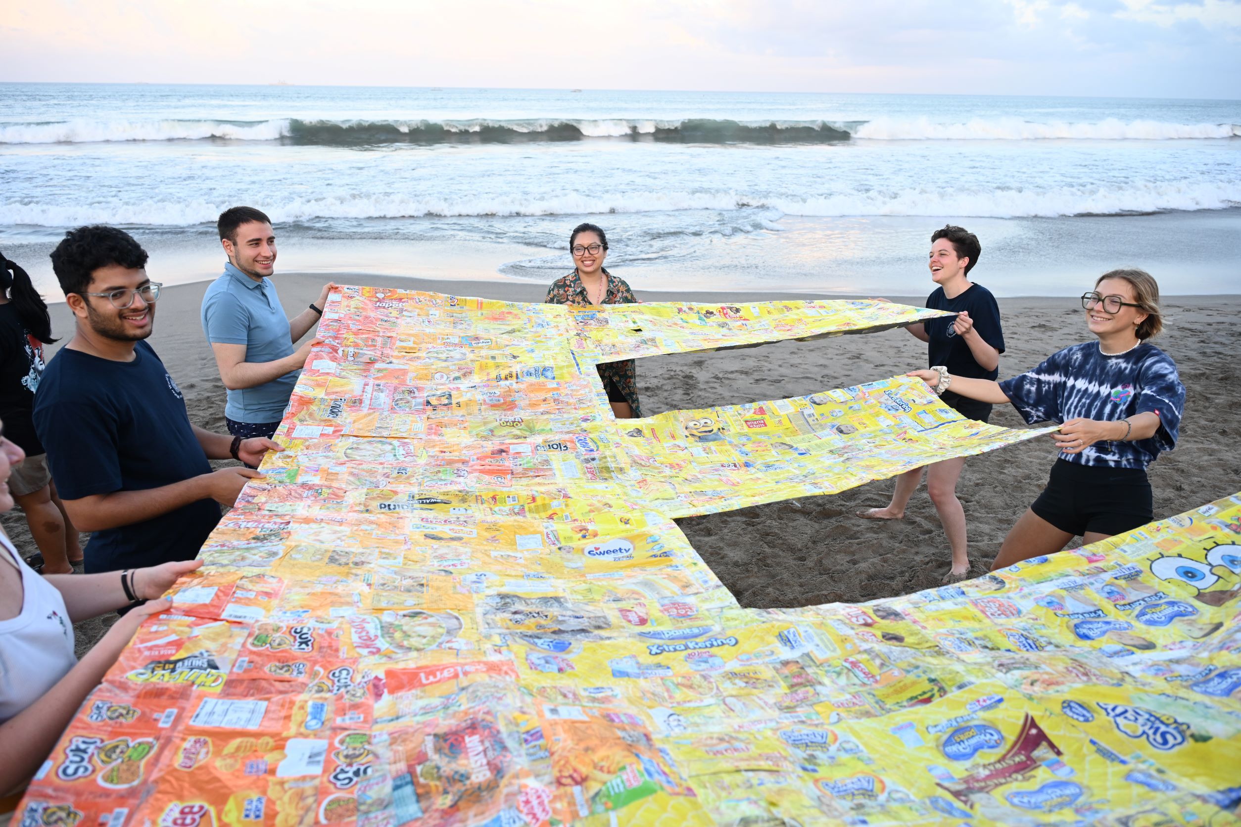 5 Jugendliche stehen an einem Strand, mit dem Ozean im Hintergrund. Gemeinsam halten sie einen riesigen Buchstaben 'E', der aus Plastikabfällen hergestellt wurde. Der Buchstabe ist gelb.