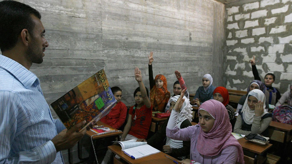 Syrische Flüchtlingsmädchen sitzen in einem Klassenzimmer im Libanon und strecken die Hand.