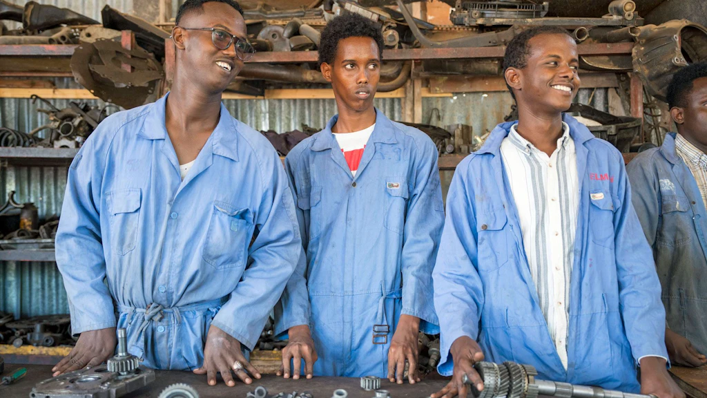 Drei junge Männer in blauen Overalls lehnen an eine Werkbank, auf der mehrere Werkzeuge liegen.
