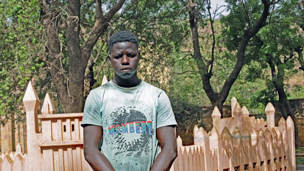 Mamadou trägt ein helles T-Shirt und steht vor einem Zaun.  