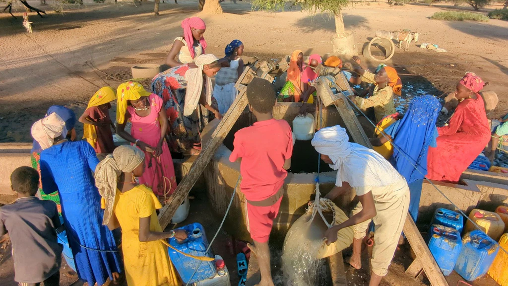 Frauen und Kinder in bunten Kleidern stehen um einen Brunnen. Ein Mann giesst einen Eimer Wasser aus.