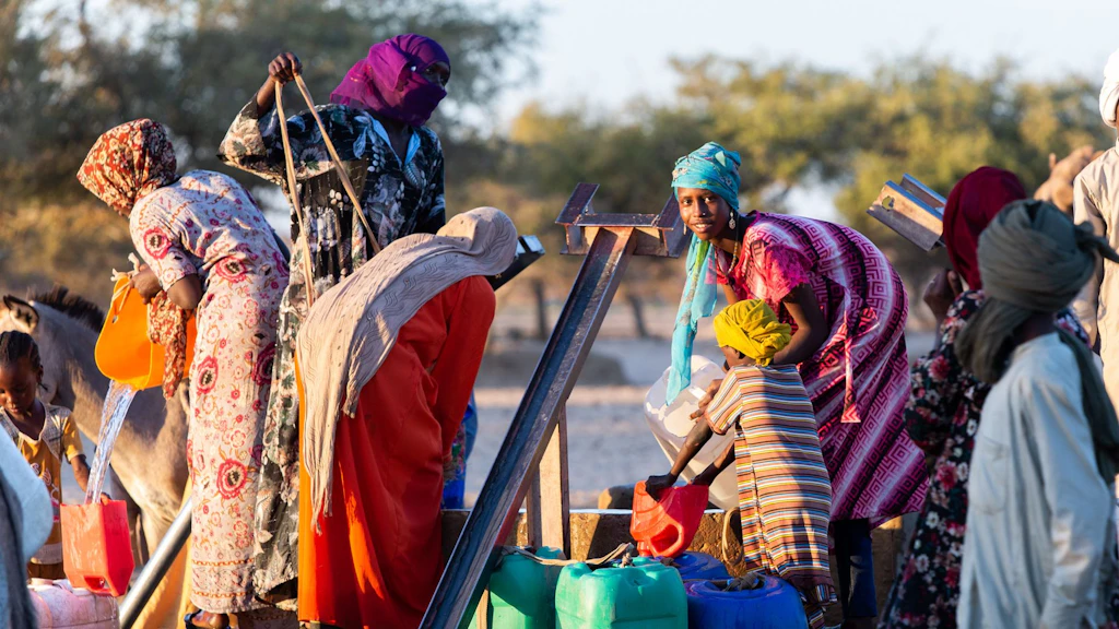 Mehrere Frauen in bunten Kleidern und Kopftüchern stehen mit Kindern am Dorfbrunnen. Sie schöpfen Wasser in verschiedenfarbige Kanister.