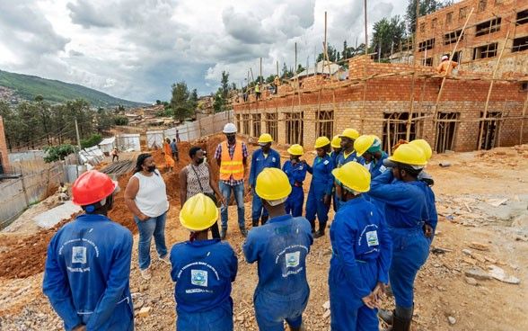 Eine Frau spricht mit einer Gruppe, die im Halbkreis steht und allesamt in einem blauen Overall gekleidet sind. Im Hintergrund bauen Arbeiter und Arbeiterinnen ein Haus aus Ziegelsteinen.