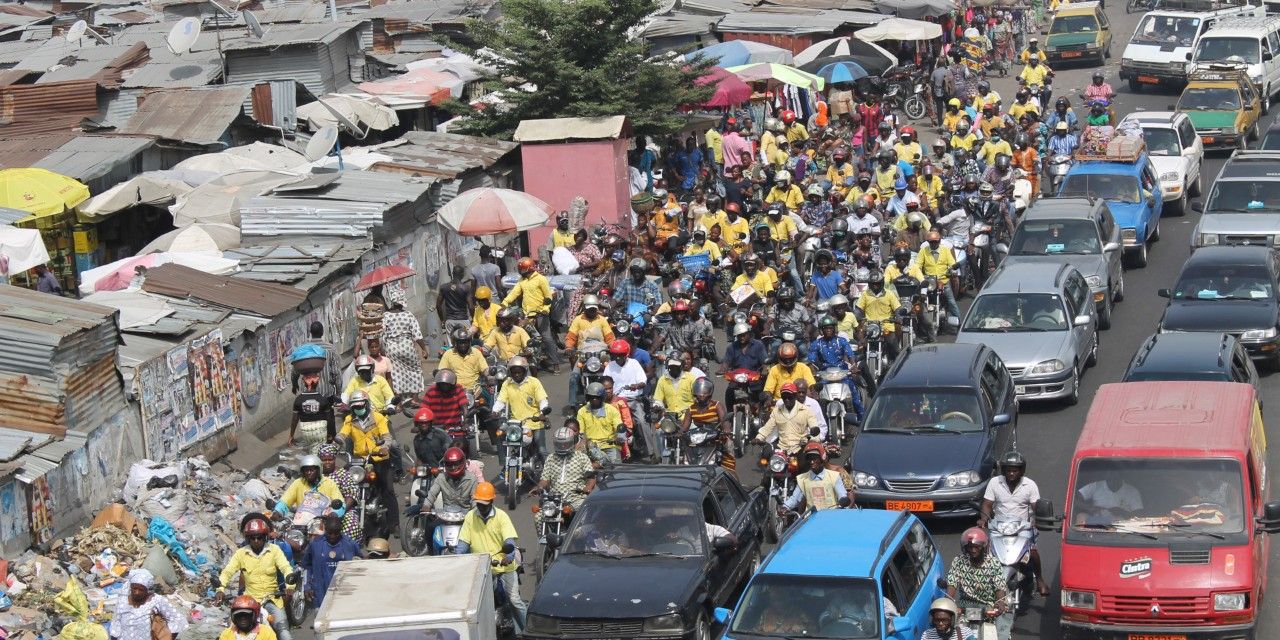 Autos und Motorräder fahren auf einer Strasse in Benin. Entlang der Strasse wurden informelle Siedlungen errichtet.