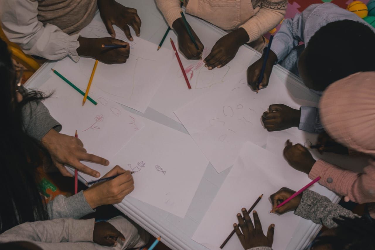 Children are bent over and drawing around a white table containing sheets of paper and coloured pencils.