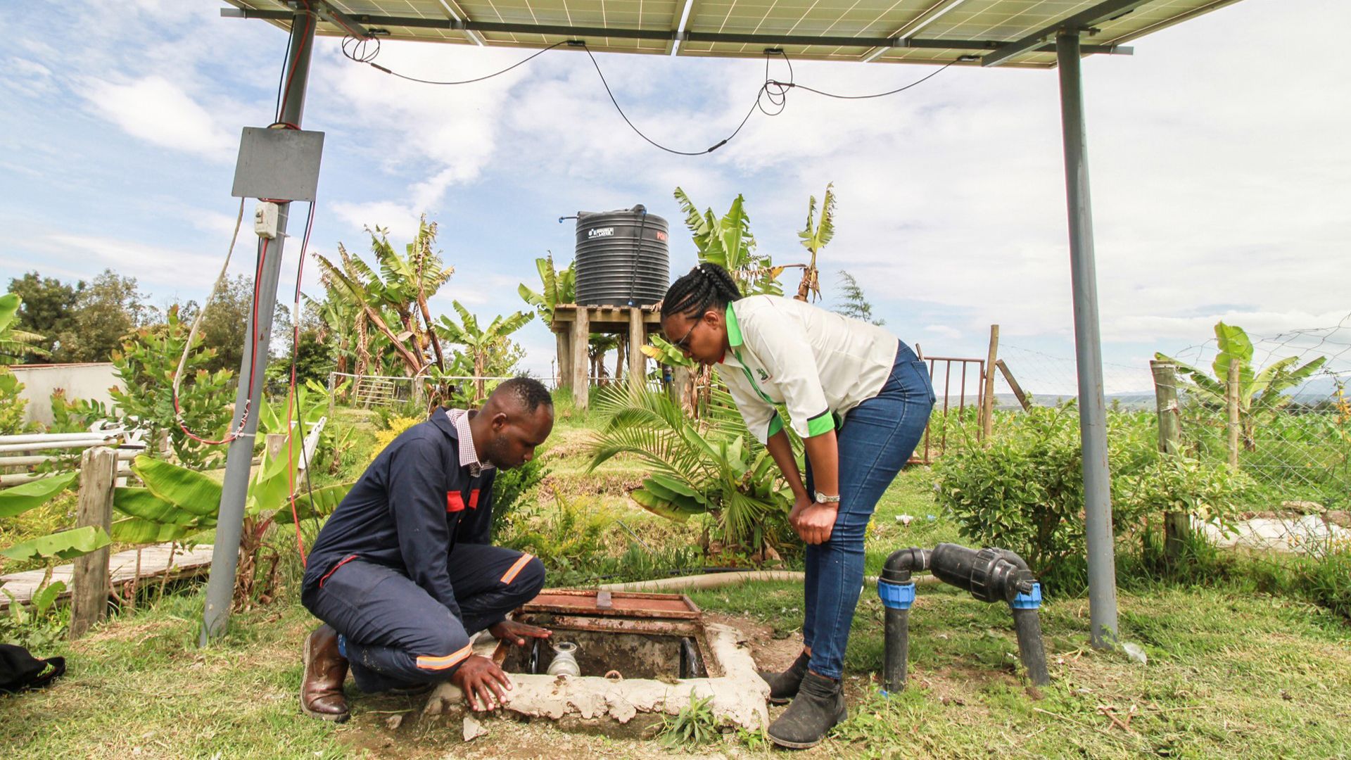 Two people inspect a solar-powered water pump system under a solar panel structure in a lush, green environment with a water tank and banana plants in the background.