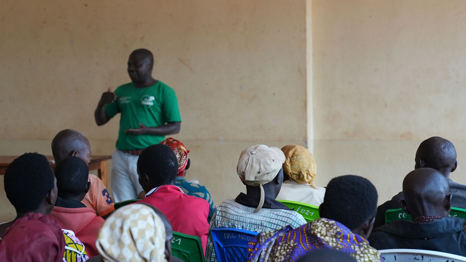 A man wearing a green T-shirt and white trousers standing and speaking in front of men and women seated on chairs of different colours.