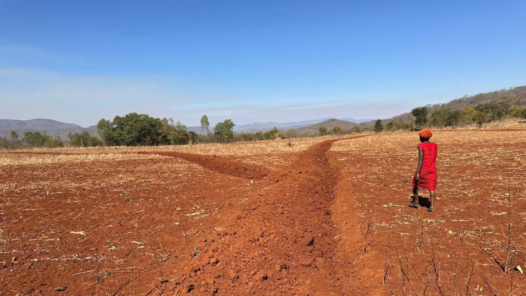 A person in a red dress walks across a dry, reddish-brown field with a trench for water harvesting. Trees and hills are visible in the background under a clear blue sky.