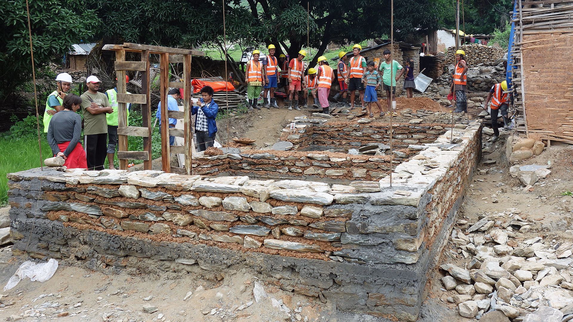 The image shows many people standing around the foundations of a house. They are wearing protective helmets and high-visibility vests.