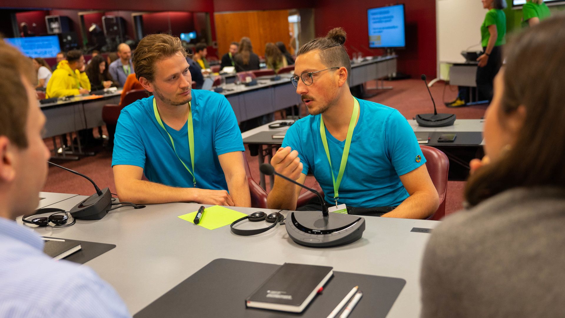Two seated men in blue T-shirts are talking.