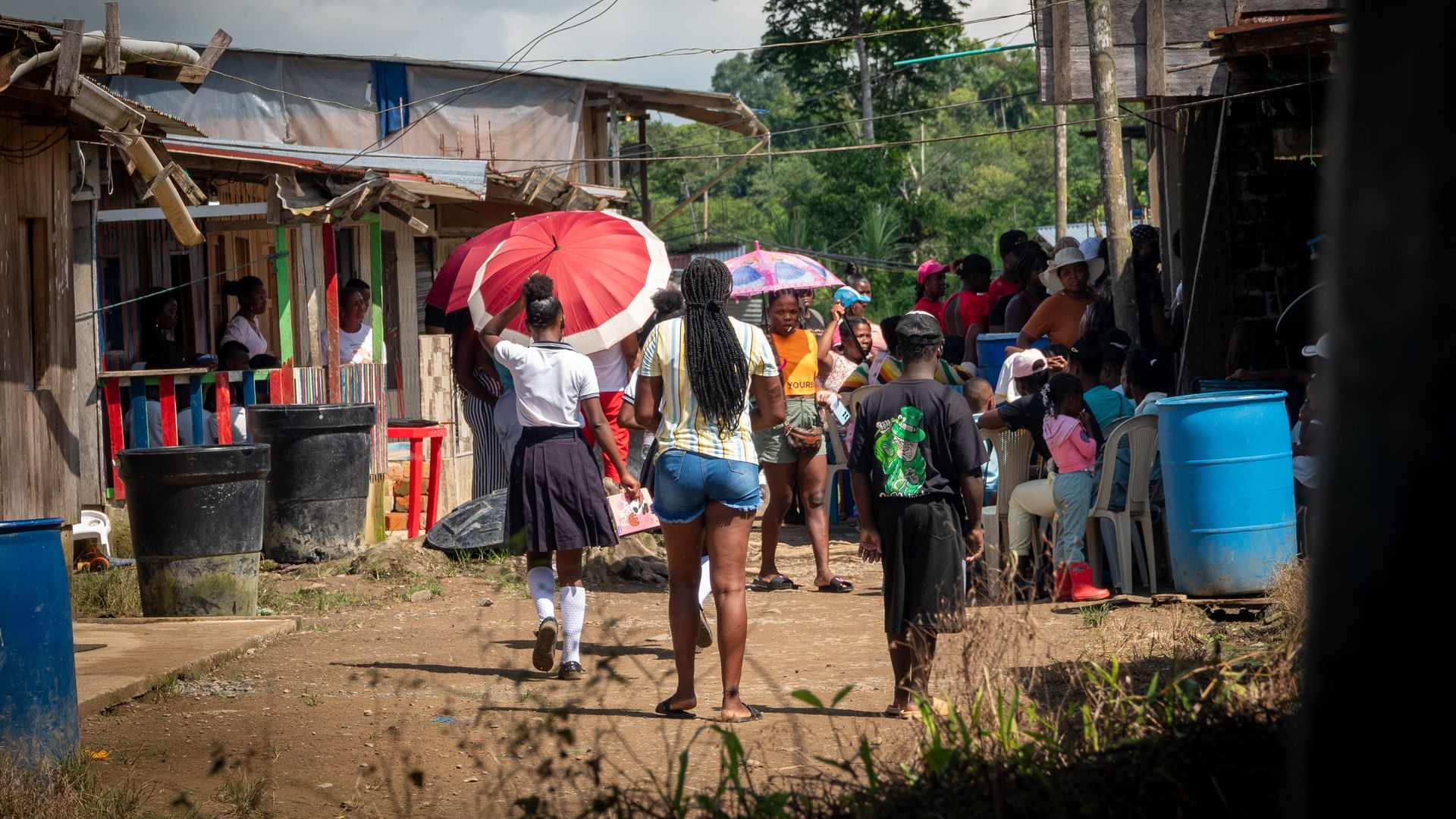 Lively village scene. People stroll along a street, some holding colourful umbrellas, with green trees in the background.
