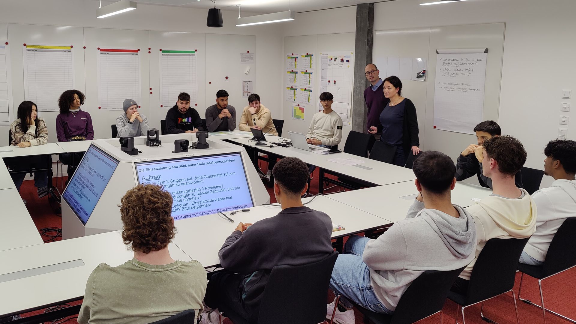 A school class around a U-shaped seating arrangement in a meeting room with monitors. Two experts in front explain the problem.