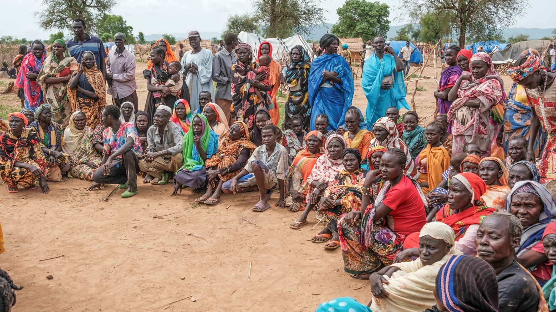 Several dozen people – men, women and children – are standing up or sitting down directly on the dirt floor. 