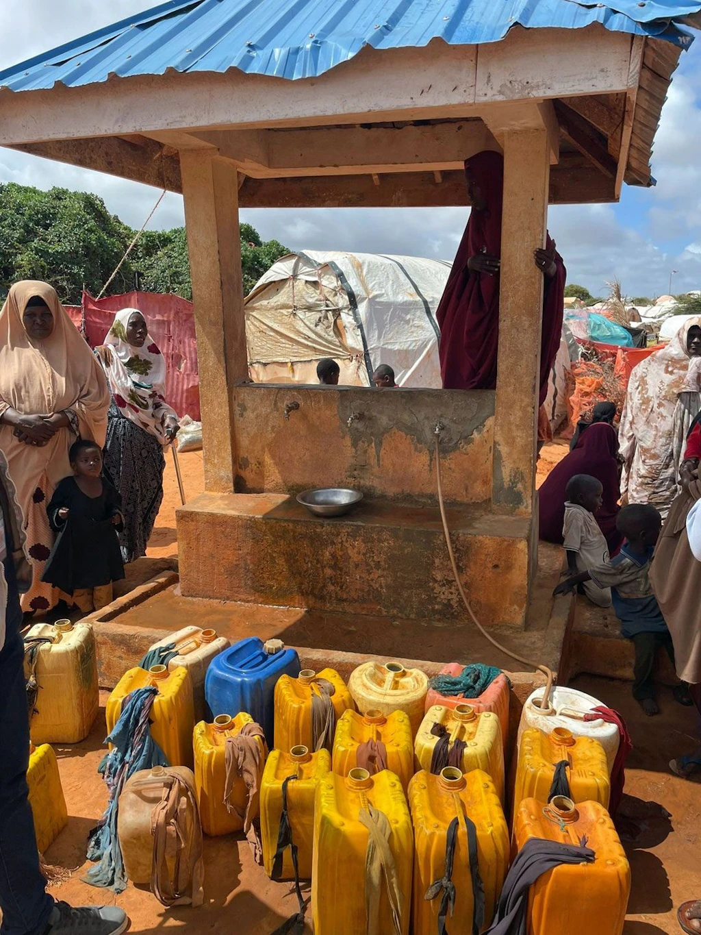 A group of women accompanied by children are standing next to a stone shelter used as a water point. Orange jerrycans can be seen in the image.