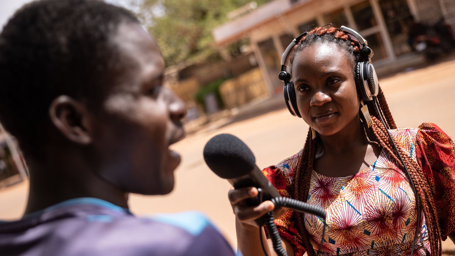 A woman in a bright dress wearing headphones holds up a microphone to a man she is interviewing on the street.