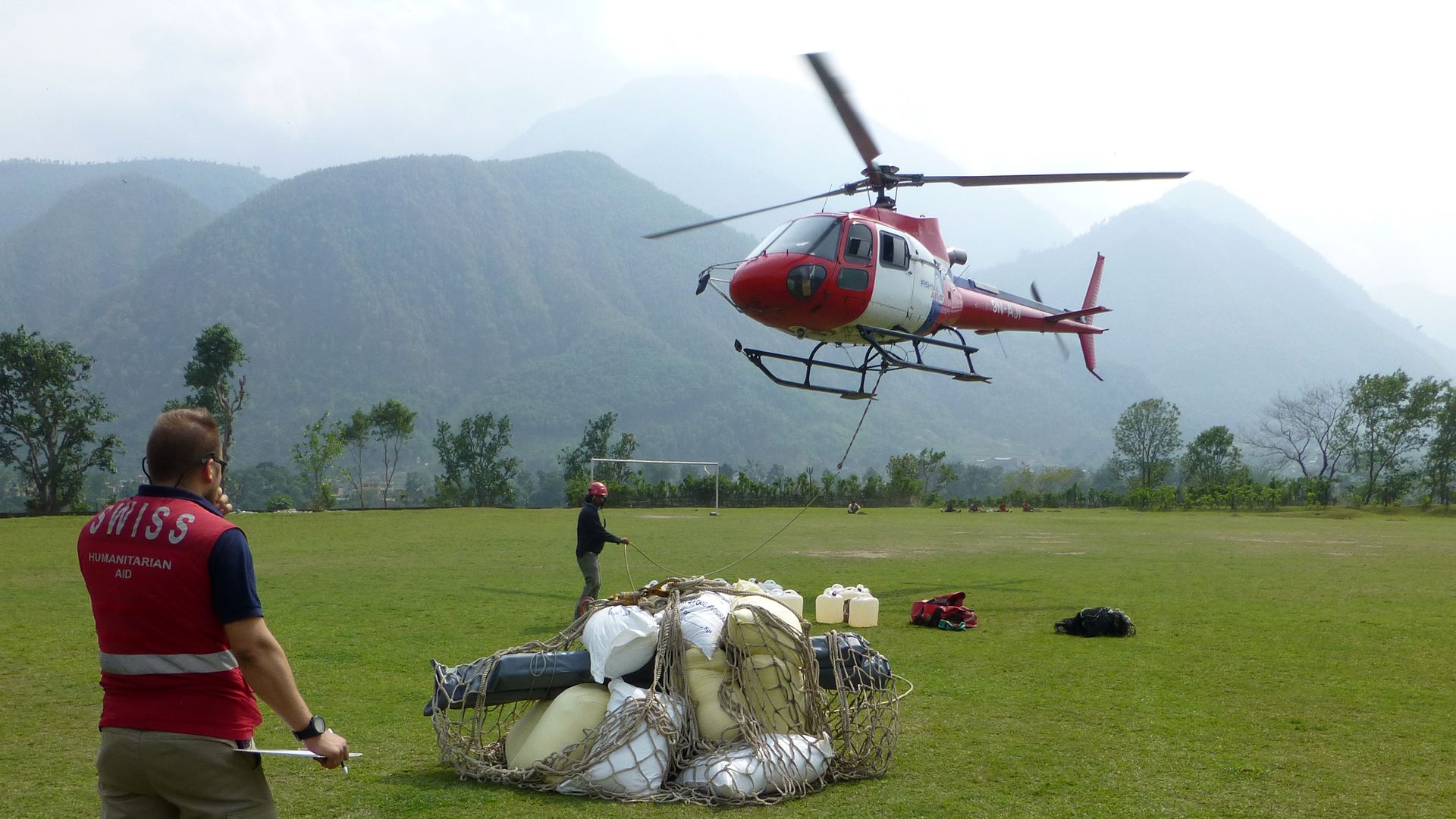 The image shows a man wearing a Swiss Humanitarian Aid vest, with a net in front of him containing relief supplies (white packages and containers). The Nepalese mountains can be seen in the background and an airborne Swiss helicopter in the foreground.