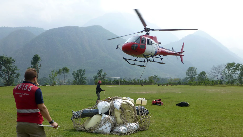 The image shows a man wearing a Swiss Humanitarian Aid vest, with a net in front of him containing relief supplies (white packages and containers). The Nepalese mountains can be seen in the background and an airborne Swiss helicopter in the foreground.