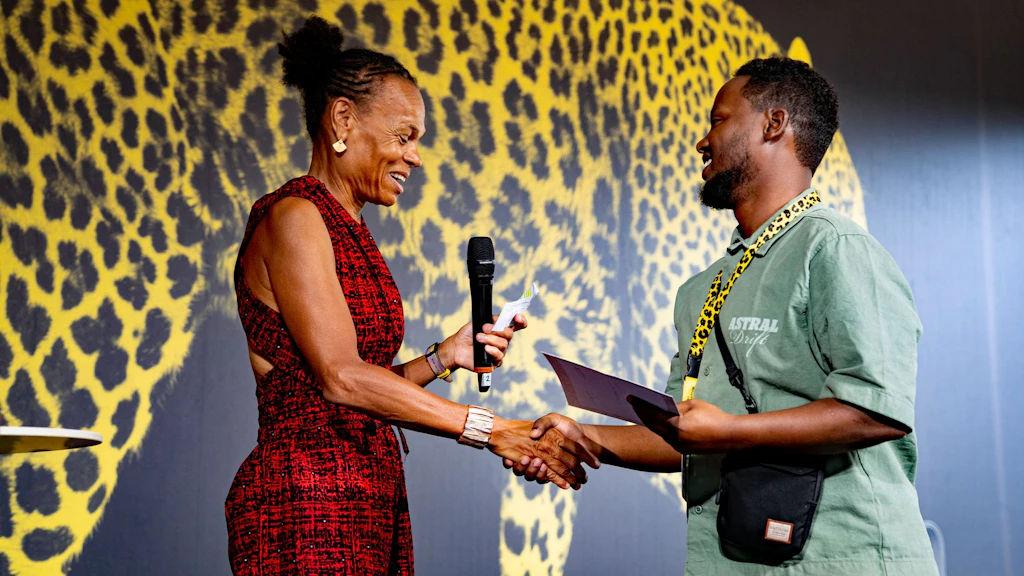 Patricia Danzi is holding a microphone and presenting an award to Mohammed Shiekh on stage at Open Doors 2026. 