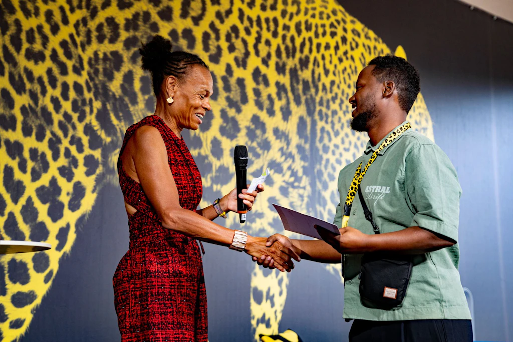 Patricia Danzi is holding a microphone and presenting an award to Mohammed Shiekh on stage at Open Doors 2026. 