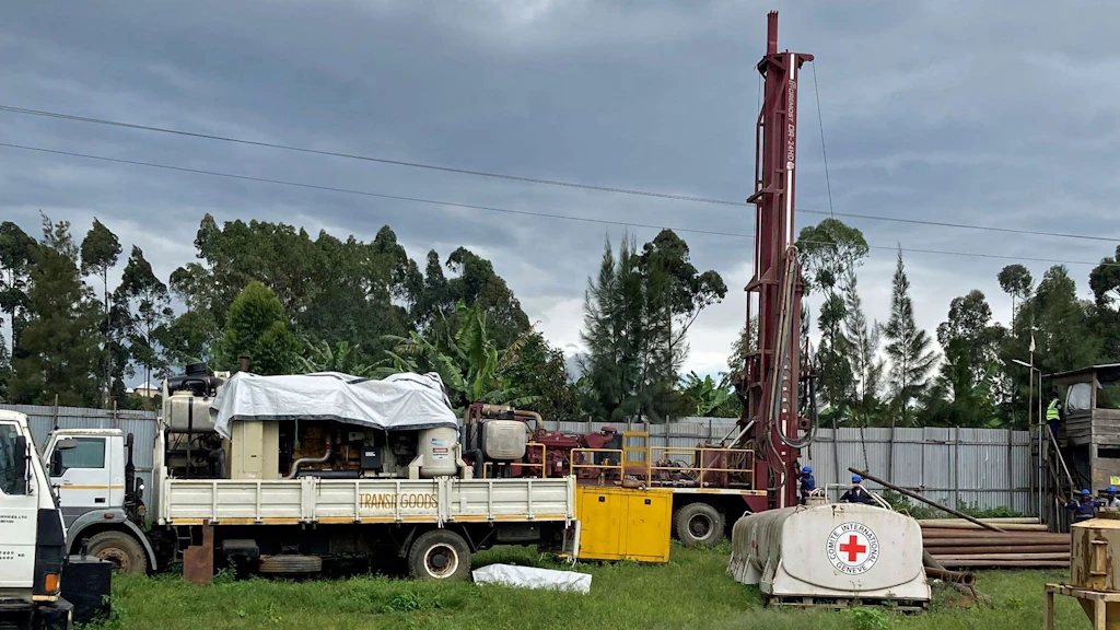 A white trailer carrying drilling equipment is parked on a grassy area with the ICRC emblem visible on a nearby machine.