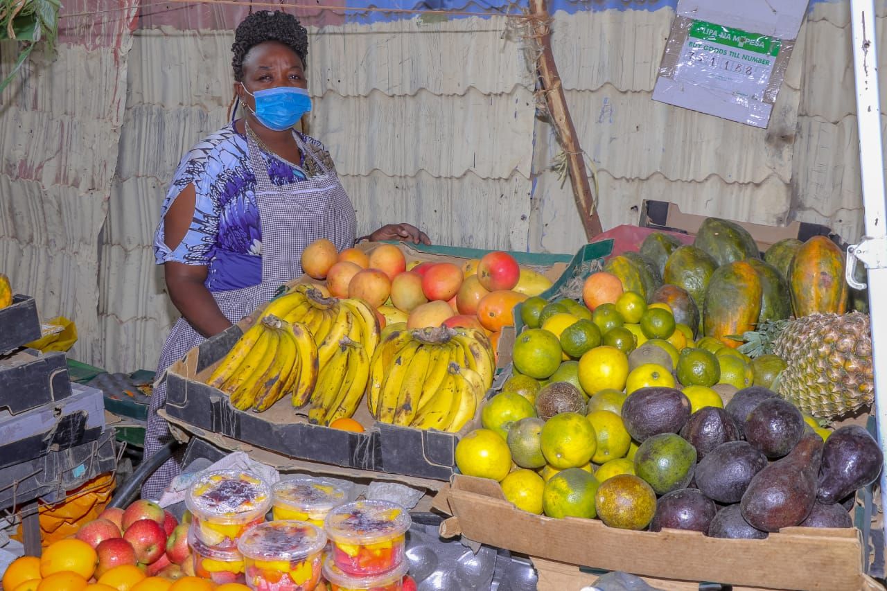 A woman wearing a blue protective mask stands to the side behind her fruit stand. Various fruits are on offer: bananas, citrus fruits, pineapples, apples, papayas.