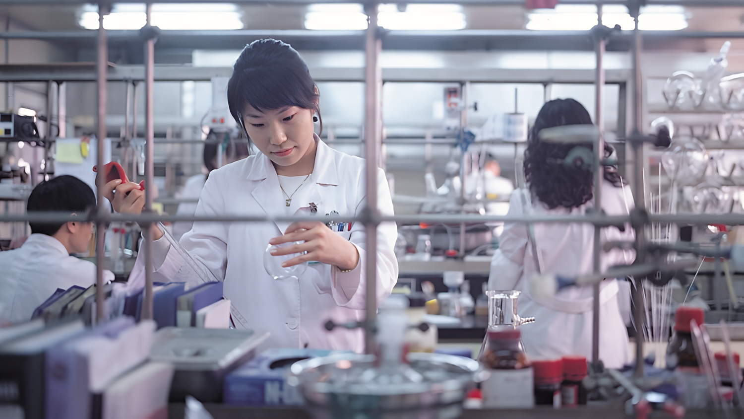 A female scientist is taking measurements in a medical laboratory. In the background, a man and a woman are also working with scientific equipment.