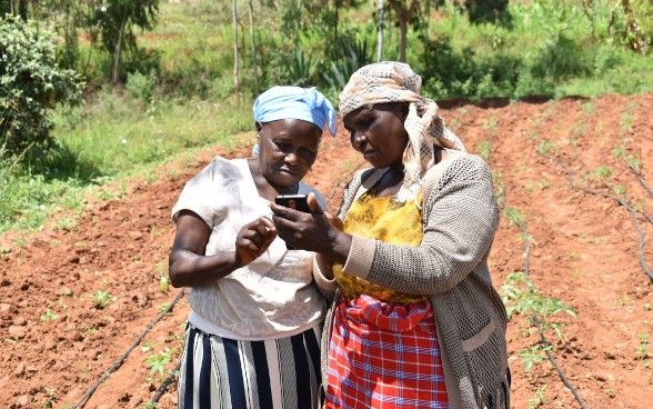 Two women are standing on a field. The woman on the right is holding a phone in her left hand. Both women are looking at the phone.