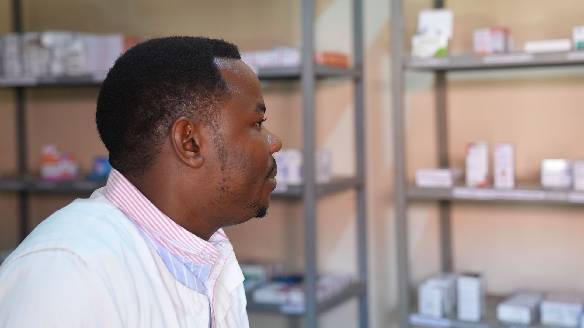 A healthcare worker, wearing a white coat over a shirt with thin red stripes, looking towards a shelf containing boxes of medicines.