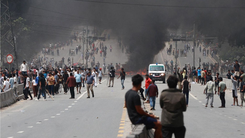 People are standing in groups on the street, with a barricade burning in the background, sending up black smoke.  