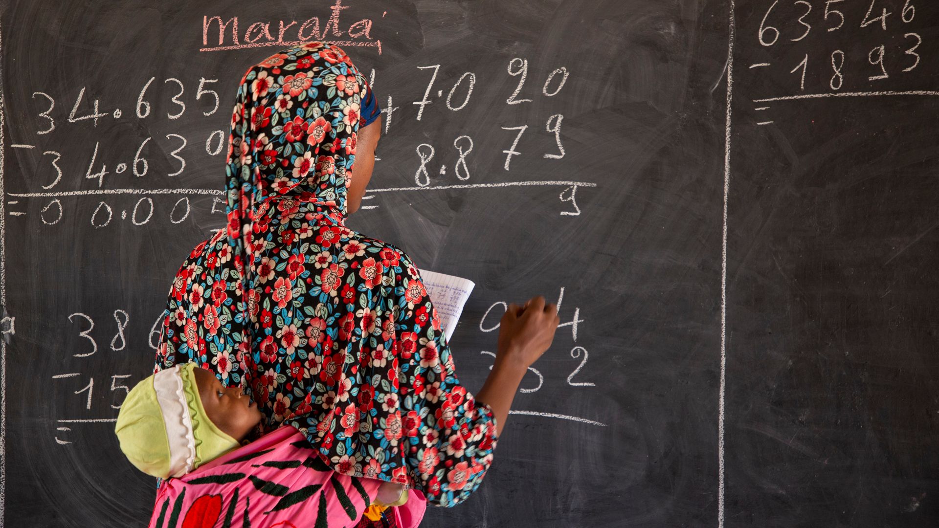 A woman stands with her back to the camera and writes numbers on a blackboard. She is carrying a small child on her back.